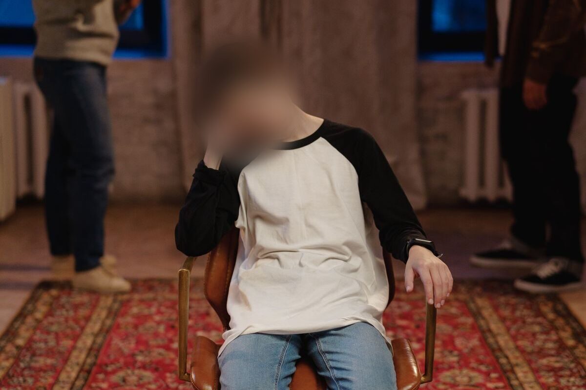 Niño con el rostro difuminado, sentado en una silla mientras los adultos hablan al fondo